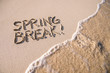 Excited Spring Break message handwritten on the smooth sand of an empty beach with an oncoming wave