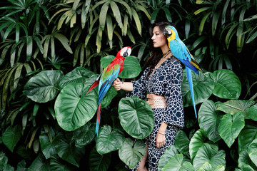 Portrait of young attractive brunette  woman with ara parrots on her hand on tropical background.