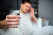 © estradaanton - Young man in bed. He hold glass of water and show it to camera. Model hold hand on forehead. Headache or hangover.