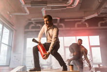 Scared Businessman Screaming While Holding Extinguisher And Standing On Desk Near Coworkers In Office With Smoke