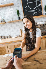 attractive cashier standing near bar counter in brown apron and holding terminal wile man making transaction in coffee house