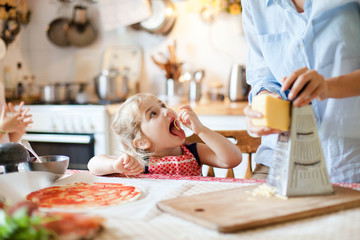 Wall Mural - Family cooking pizza in kitchen. Mother and daughter preparing homemade italian food. Funny little girl gourmet is helping woman, eating and tasting cheese and ingredients. Children chef concept.