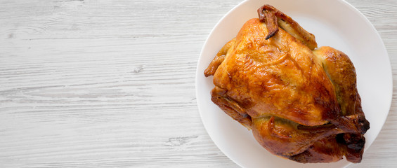 Homemade tasty rotisserie chicken on white plate over white wooden background, top view. Copy space.