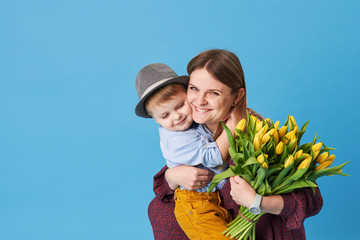 Young mother hugs his little son sitting on the floor against a blue background. Nearby lies a bouquet of spring yellow flowers. Care and relationships and family concept. Mothers day