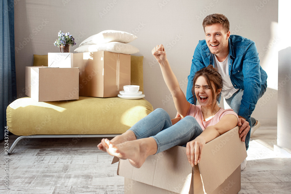 Young family couple bought or rented their first small apartment. Cheerful woman scream sitting in box. Guy move her. They play game during moving in. - obrazy, fototapety, plakaty 