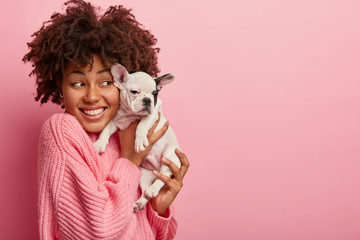 Indoor shot of happy dark skinned lady with pleased facial expression, wears pink oversized jumper, holds little puppy closely to face, isolated over rosy background, mock up space, sincere emotions