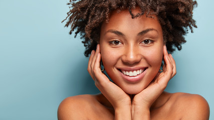 Wall Mural - Close up portrait of relaxed black woman has gentle skin after taking shower, satisfied with new lotion, has no makeup, smiles tenderly, shows perfect teeth, stands shirtless against blue background