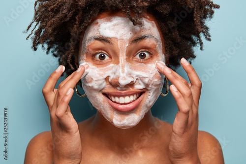 Smiling female massages face, washes with soap, demonstrates cleanliness or softness of skin, has curly haircut, models over blue background, has naked body, beautiful smile, bubble facial foam
