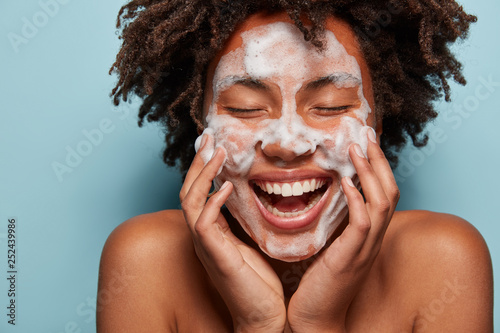 People, skin care and rejuvenation concept. Pleased dark skinned woman with soft skin, applies white foam on face, touches cheeks, smiles broadly, cares of hygiene, feels relaxed and satisfied