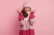 Isolated shot of overjoyed woman raises hands, wears knitted hat and loose sweater, expresses positive emotions, laughs sincerely at funny story, isolated over pink background. Emotions concept