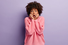 Neurotic African American Woman Looks Stressfully At Camera, Keeps Both Hands Near Mouth, Scared Of Fearful Thing, Dressed In Pink Oversized Jumper, Isolated Over Purple Background. Omg Concept