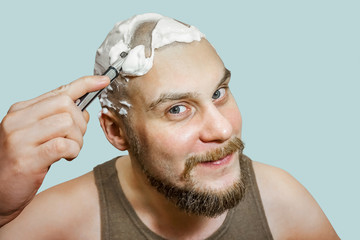 A guy with a beard and a razor in his hand, with foam and gel on his face shaves in the morning at home on a colored background