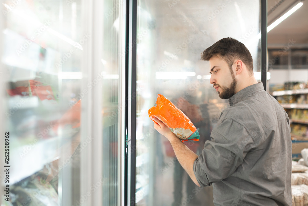 Buyer standing in the supermarket near the refrigerator holds the package in his hand and reads the label. Man checks the date of production of frozen food in the supermarket. - obrazy, fototapety, plakaty 
