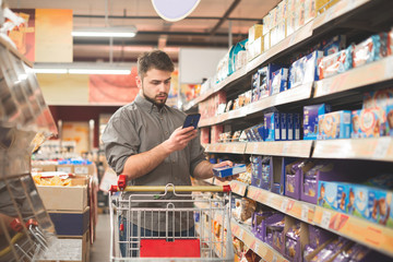 portrait of a man standing with a cart in the department of sweets in a supermarket, using a smartph