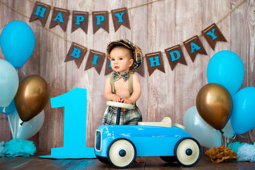 Little boy kid gentleman in retro costume with suspenders and cap is sitting on a wooden car. Children's party with balloons Happy Birthday, 1 year