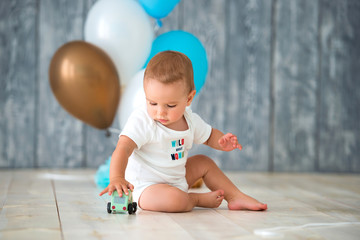 Cute little boy 1 year old sits on a warm wooden floor and plays with a toy car. Behind the plan birthday balloons