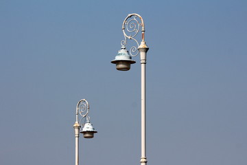 Two baroque style light grey public lights mounted on rusted wrought iron decorated poles on clear blue sky background