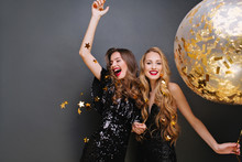 Brightfull Expressions Of Happy Emotions Of Two Amazing Girls Celebrating Party On Black Background. Luxury Black Dresses, Smiling, Golden Tinsels, Big Balloon, Long Curly Hair, Stylish Look