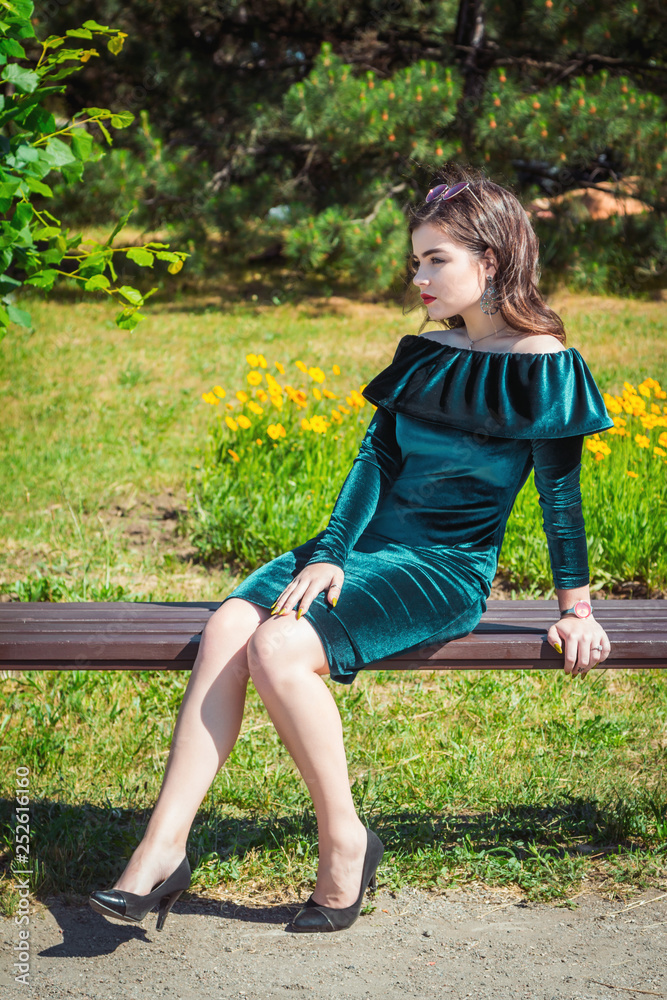 Portrait of beautiful young brunette woman with bright makeup, wearing emerald  green velvet dress, sitting on a bench in a park, with yellow flowers and  trees on background Stock Photo | Adobe
