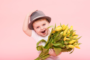 Adorable smiling child with spring flower bouquet looking at camera isolated on pink. Little toddler boy in hat holding yellow tulips as gift for mom