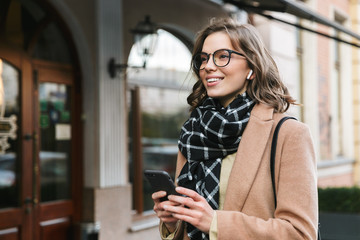 Wall Mural - Young woman outdoors walking by street using mobile phone listening music with earphones.