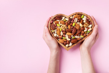 Wall Mural - Womans hands holding heart shaped bowl with mixed nuts on pink table top view. Healthy food and snack. Flat lay.