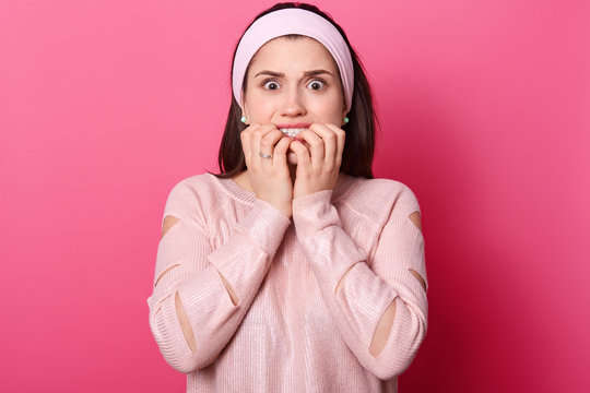 Woman with both hands in front of mouth. Scared girl in pink sweater and hairband, isolated over rose background. Lady wathes horror film alone. Female afraids of something. People concept.