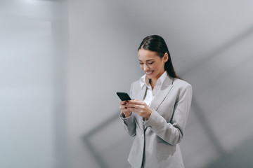 Wall Mural - Smiling attractive brunette using smart phone in formal wear standing in hall and writing e-mail on smart phone. Work hard, dream big, never give up.