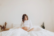 Happy Pregnant Woman In White Holding Belly Bump And Relaxing On White Bed At Home. Stylish Pregnant Mom Waiting For Baby. Motherhood And Fertility Concept. Maternity Time