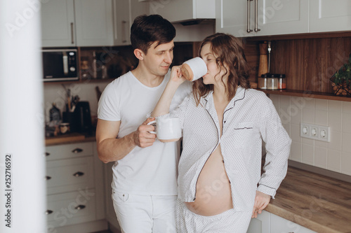 Happy Young Pregnant Couple In White Pajamas Holding Cups And