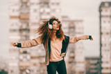 Fototapeta  - Beautiful mixed race hipster teenage girl with curly hair enjoying music and jumping. In background blurred buildings.