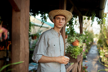 Wall Mural - Guy gardener in a straw hat is standing with plastic glass in his hand next to a wooden veranda in the wonderful nursery-garden on a sunny day.