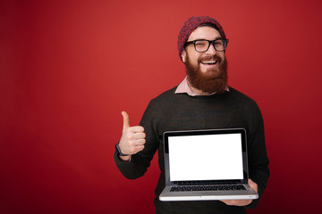 Wall Mural - Smiling bearded man in eyeglasses which showing blank laptop screen and thumb up while looking at camera. Isolated red background