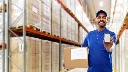 Poster - mail service, technology and shipment concept - happy indian delivery man or warehouse worker with smartphone and parcel box in blue uniform over goods background