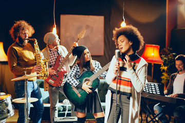 Canvas Print - Mixed race woman singing. In background band playing instruments. Home studio interior.