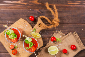 Two glasses of tomato juice decorated with fresh tomatoes, cucumber and leaves on a wooden background