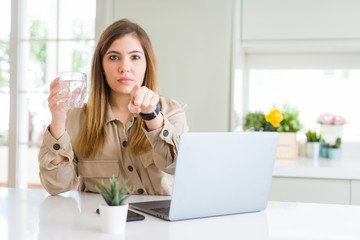 Wall Mural - Beautiful young woman working with computer takes a break to drink glass of water pointing with finger to the camera and to you, hand sign, positive and confident gesture from the front