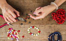 Making Bracelet Of Colorful Beads. Female Hands With A Tool On A Rough Wooden Table.