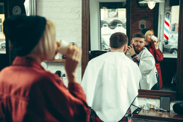 Wall Mural - Client during beard shaving in barbershop. Female barber at salon. Gender equality. Woman in the male profession.