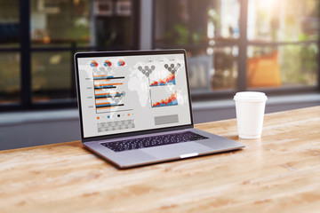 Close up of laptop with graphs, charts on screen,cup of coffee on table in empty room, cafe. Computer on wooden desk.