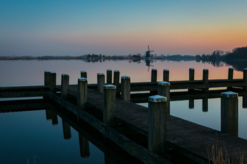 Wall Mural - a wooden pier near a calm lake close to rotterdam, holland at sunset. a windmill is visible in the d