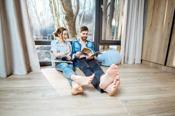 Happy couple sitting together on the floor near the window with beautiful view, reading some magazines and relaxing at home