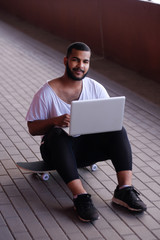 Wall Mural - Smiling African-American guy is sitting on a bridge sidewalk, holding the laptop on his knees and looking at a camera