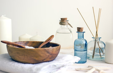 On a wooden white table transparent bottles of white and blue with corks, inside cosmetics for skin care. Near incense sticks in containers, white candles and a wooden cup with a spoon, home spa