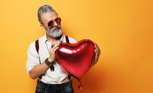 Senior Bearded Old Man In Aviator Sunglasses Holds Gently Red Heart Balloon For Valentines Day On Yellow Background