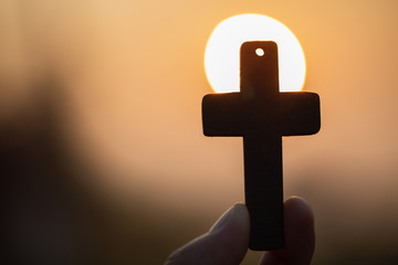 Woman hand holding holy lift of christian cross with light sunset background.