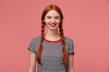 Wall Mural - Nice pretty red-haired girl with red lips, two braids, chrarming smile, shows white healthy teeth, dressed in stripped t-shirt, isolated on a pink background