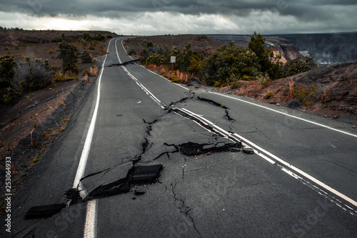 Fotomural Damaged asphalt road (Crater Rim Drive) in the Hawaii Volcanoes National Park after earthquake and eruption of Kilauea (fume at upper right) volcano in May 2018