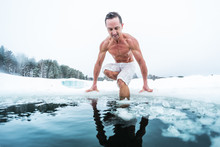 Young Man With Lean Muscular Body Going To Swim In The Cold Winter Water With Ice Floating On The Surface And Forest On The Background