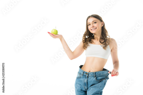 Fotomural smiling slim young woman in big jeans holding green apple isolated on white